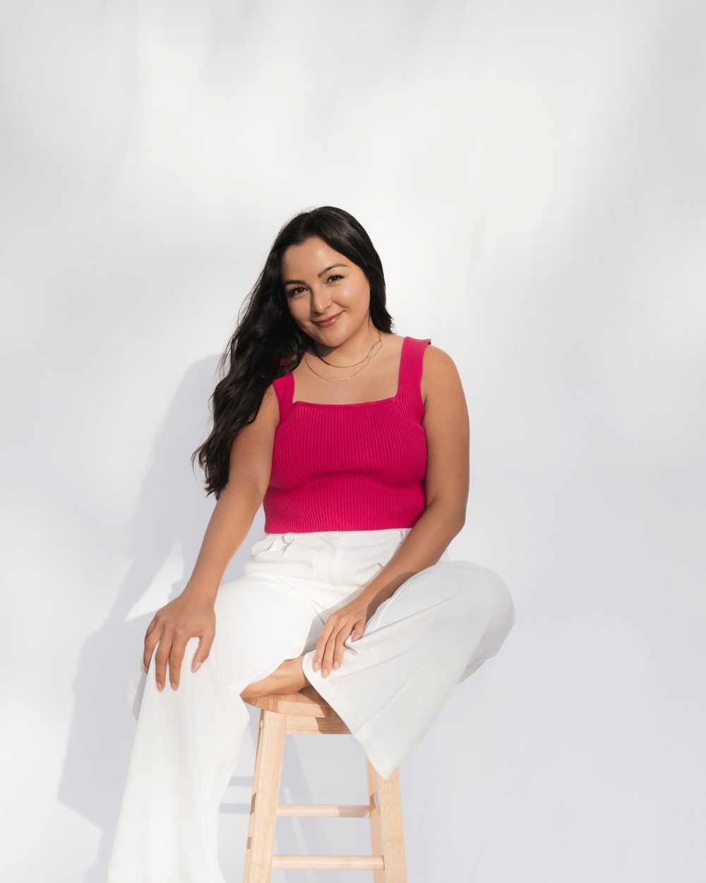 Sarah Ortega smiling, seated on a wooden stool in a pink top and white trousers